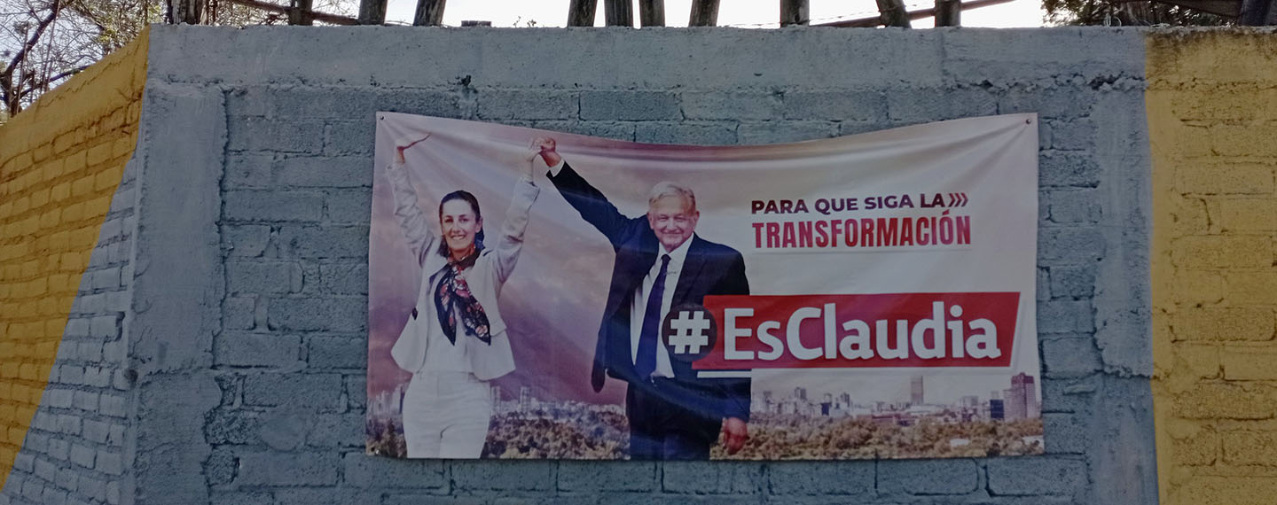 Claudia Sheinbaum y Andrés Manuel López Obrador en un cartel de propaganda electoral de MORENA en Dolores Hidalgo, Guanajuato (México). El cartel está colgado en una pared de bloques de cemento pintada de gris y amarillo. En la parte superior de la pared sobresalen trozos de madera con una alambrada